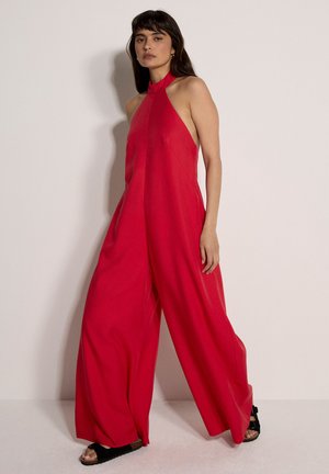 Donna che indossa una tuta rossa smanicata a gamba larga con scollo alto, in piedi contro uno sfondo chiaro e semplice, indossando sandali neri.