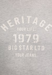 Pelēks džemperis ar baltu, nolietotu tekstu: "MANTOJIET SAVU DZĪVI. 1979 BIGSTAR LTD JŪSU DŽINSI." Klasisks apaļais izgriezums, maiga tekstūra.