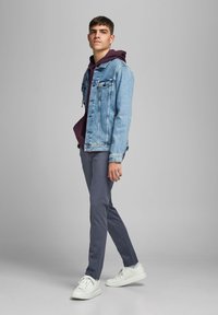 Chaqueta de mezclilla azul claro, sudadera color burdeos debajo, pantalones grises y zapatillas blancas. Mostrado en una pose de pie casual contra un fondo neutro.