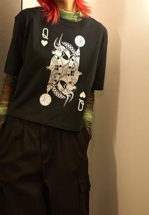 Person mit roten Haaren trägt ein schwarzes, kurz geschnittenes T-Shirt mit einem Motiv der Herz-Dame, die Basketbälle hält, darüber grün gemusterte Langarmärmel.