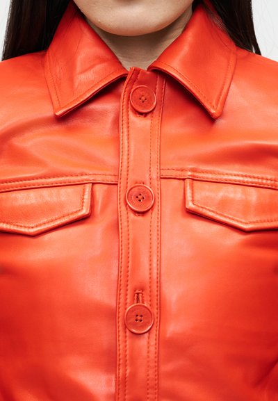 Κοντινό πλάνο ενός ατόμου που φορά ένα φωτεινό πορτοκαλί δερμάτινο μπουφάν με μεγάλα κουμπιά και δύο εμπρόσθιες θήκες με καπάκι.