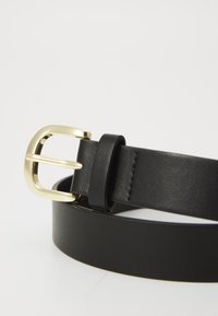 Ceinture en cuir noir avec une boucle métalliques dorée au design courbé, texture lisse et coutures uniformément espacées le long du bord.