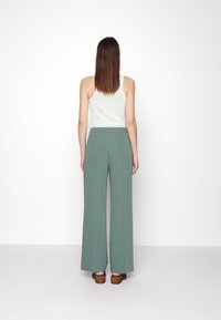 Pantalons à jambe large en tissu vert doux, présentant une taille haute, une texture lisse et associés à des sandales à talons en bois marron.