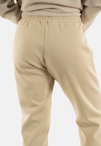 Pantalones de chándal beige con una cintura elástica y un corte relajado. Textura suave con costuras mínimas y un pequeño logo cerca de la cintura.
