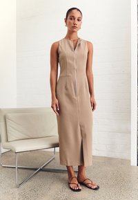 Vestido midi sin mangas en color beige con cremallera frontal, bolsillos laterales y un corte recto. Combinado con sandalias planas negras. Diseño minimalista.