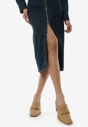 Denim dress with a front zipper, knee-high slit, and fitted design. Beige heeled loafers with a bow accent.
