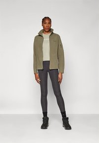 Giacca in pile verde oliva con collo alto, tasche con cerniera, abbinata a una t-shirt grigio chiaro e leggings neri, stivali da passeggio con suola zigrinata.