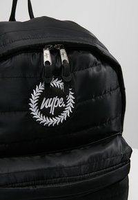 Sac à dos noir matelassé avec deux fermetures éclair argentées et un patch rond noir arborant une couronne de laurier blanche et le logo "hype.".