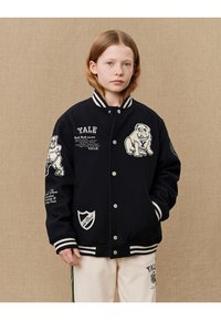 Čierna varsity bunda s bielymi pruhmi. Obsahuje vyšívané logo Yale, grafiku buldoka a textové detaily. Zapínanie na gombíky a vrecká.