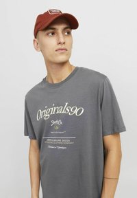 Harmaa puuvillapaita lyhyillä hihoilla, jossa on "Originals90" -logo valkoisella ja keltaisella tekstillä. Yhdistetty ruosteenväriseen lippikseen. Minimalistinen muotoilu.