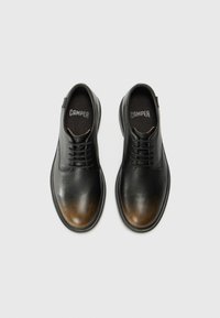 Scarpe in pelle nera con design a lacci. Presentano una texture liscia con un gradiente marrone che sfuma verso la punta. Logo "Camper" visibile all'interno.