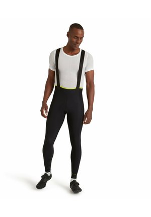 Schwarze Radsportbuxe mit schwarzen Trägern und einem neongrünen Bund, getragen über einem eng anliegenden weißen Hemd mit einem aerodynamischen Design.