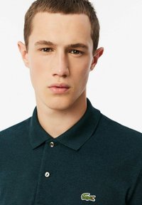 Marineblaues Poloshirt aus strukturiertem Stoff, mit einer zwei-Knopf-Leiste und einem grünen, gestickten Krokodil-Logo auf der linken Brust.