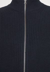 Mørk marineblå strikkegenser med høy krage og full-lengde sølvglidelås. Har et teksturert ribbestrikket design gjennom hele.