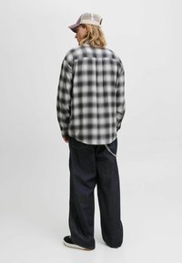 Camisa de franela a cuadros en gris y negro, de manga larga, con corte holgado, combinada con pantalones de mezclilla oscuros y zapatillas negras, con un detalle de cadena.
