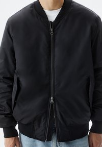 Blouson aviateur noir en matière lisse et matelassée. Il comprend une fermeture éclair à l'avant avec des éléments métalliques, des poignets côtelés et des poches latérales.