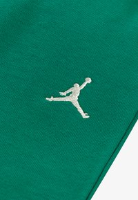 Tecido verde com uma textura suave, apresentando um logótipo Jumpman branco bordado em pleno ar, posicionado na parte inferior direita.