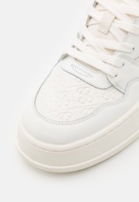 Baskets en cuir blanc avec un motif en relief, fermeture à lacets plats, col rembourré et semelle en caoutchouc distinctive. Design minimaliste avec des accents subtils.