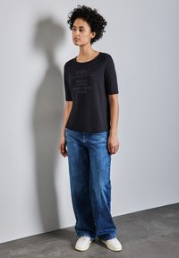 T-shirt noire à manches courtes avec du texte en police claire, associé à un jean bleu ample. Le modèle porte des baskets blanches, se tenant devant un fond gris.