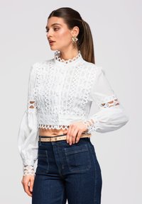 Femme portant un haut court blanc à motif de dentelle avec des manches ballon et un jean taille haute bleu foncé, posant avec la main sur la hanche.