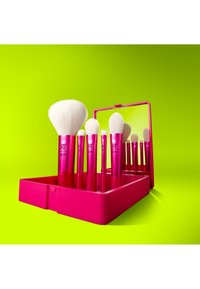 Ensemble de six pinceaux de maquillage blancs à manches roses, posés dans un étui rose assorti avec un miroir, sur un fond vert vif.