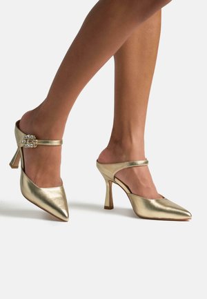 Mules de tacón alto metálicos dorados con punteras afiladas, tira en el tobillo con acento de joya, textura suave y una silueta elegante.
