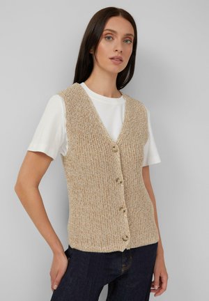 Femme portant un gilet boutonné en tricot beige sur un t-shirt à manches courtes blanc et un jean bleu foncé, devant un fond uni.
