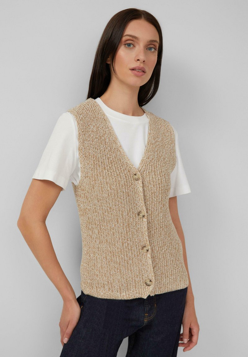 Vrouw in een beige gebreide vest met knopen over een wit T-shirt met korte mouwen en donkerblauwe jeans tegen een effen achtergrond.