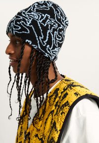 Knit beanie in black with blue abstract patterns; worn by a person with braided hair, paired with a yellow vest featuring a splatter design.