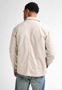 Beige cotton jacket with a collar, back seam detailing, and long sleeves, paired with light blue jeans. Clean, minimal design.