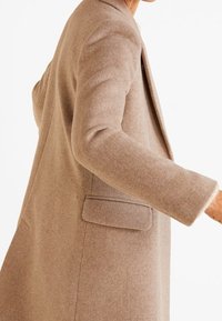 Personne portant un long manteau en laine beige avec une poche à rabat, vue de profil sur un fond blanc uni.