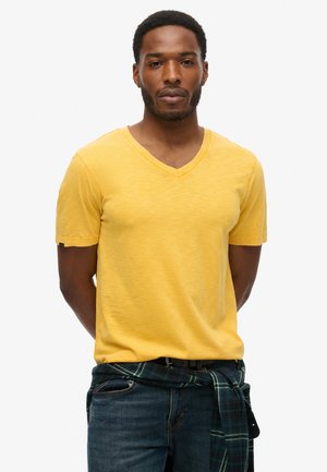 Gul v-hals t-shirt lavet af blødt stof med korte ærmer. En blå og grønternet skjorte bundet om taljen tilføjer detaljer. Denim jeans fuldender looket.