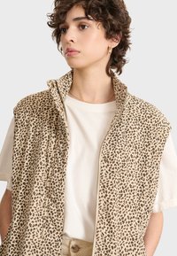 Beige väst med leopardmönster, dragkedjestängning och hög krage; bärs över en vit t-shirt som visar upp textilens struktur.