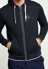 Svart zip-up hoodie i mjukt material med en frontlogotyp, vita dragsnören och kontrasterande vita kanter på ärmar och fåll.