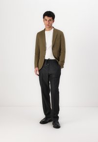Blazer verde oliva sobre una camiseta blanca, acompañado de pantalones negros a rayas y zapatos negros, con un ajuste relajado y texturas sutiles.