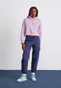 Haut de survêtement court violet clair, pantalon bleu marine à cordon de serrage et baskets teal. Texture lisse, coupe décontractée, avec logo blanc sur le haut et poignets élastiques.