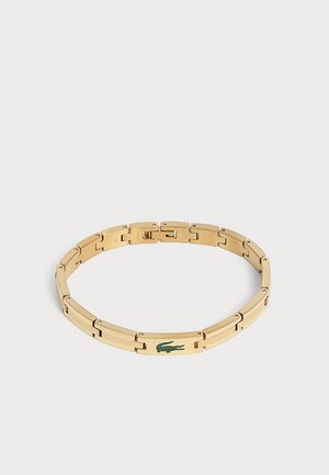 Bracelet en métal ton or avec un logo crocodile vert sur un maillon rectangulaire, présenté sur un fond clair uni.