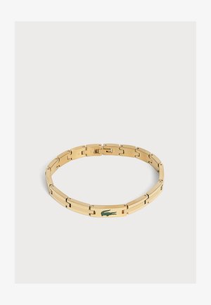 Bracelet en métal ton or avec un logo crocodile vert sur un maillon rectangulaire, présenté sur un fond clair uni.