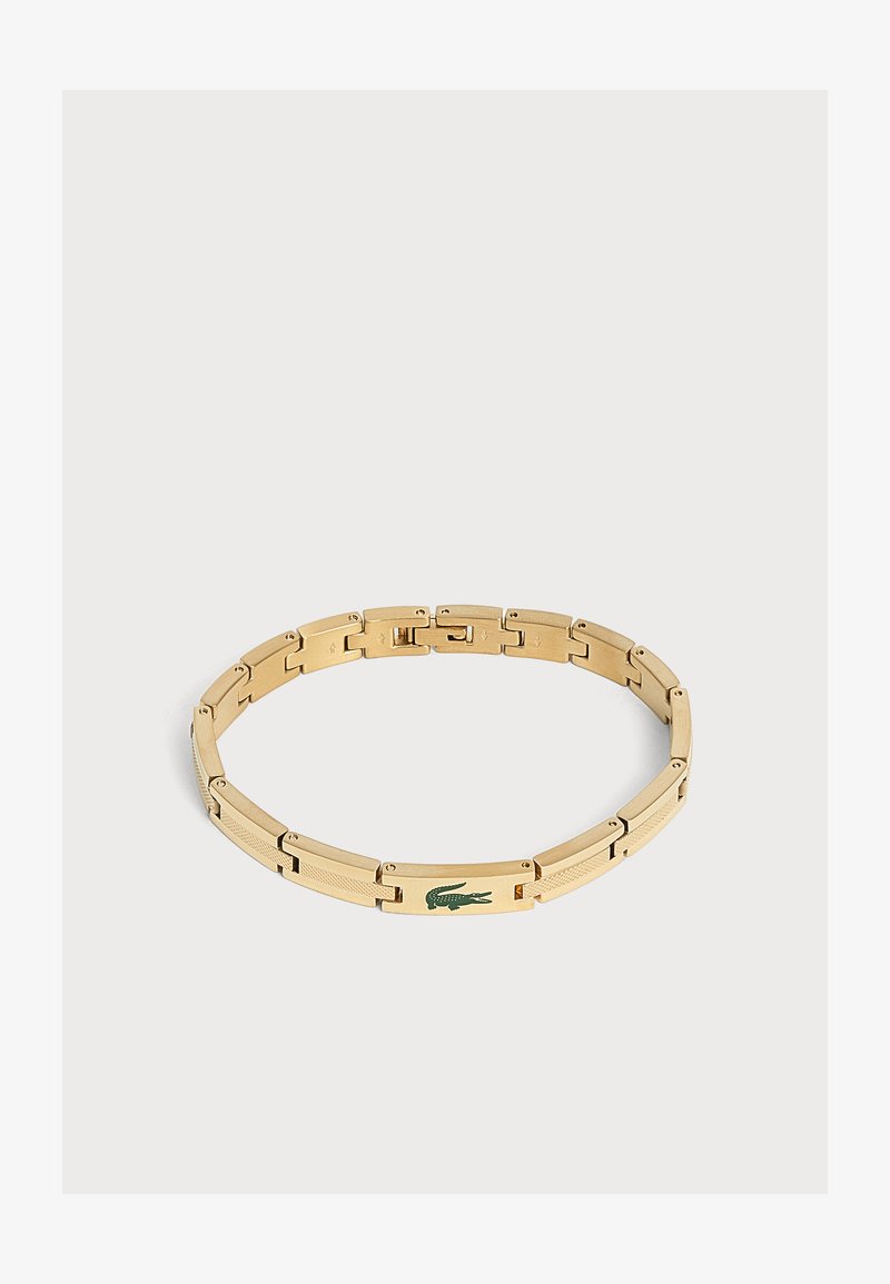 Bracelet en métal ton or avec un logo crocodile vert sur un maillon rectangulaire, présenté sur un fond clair uni.