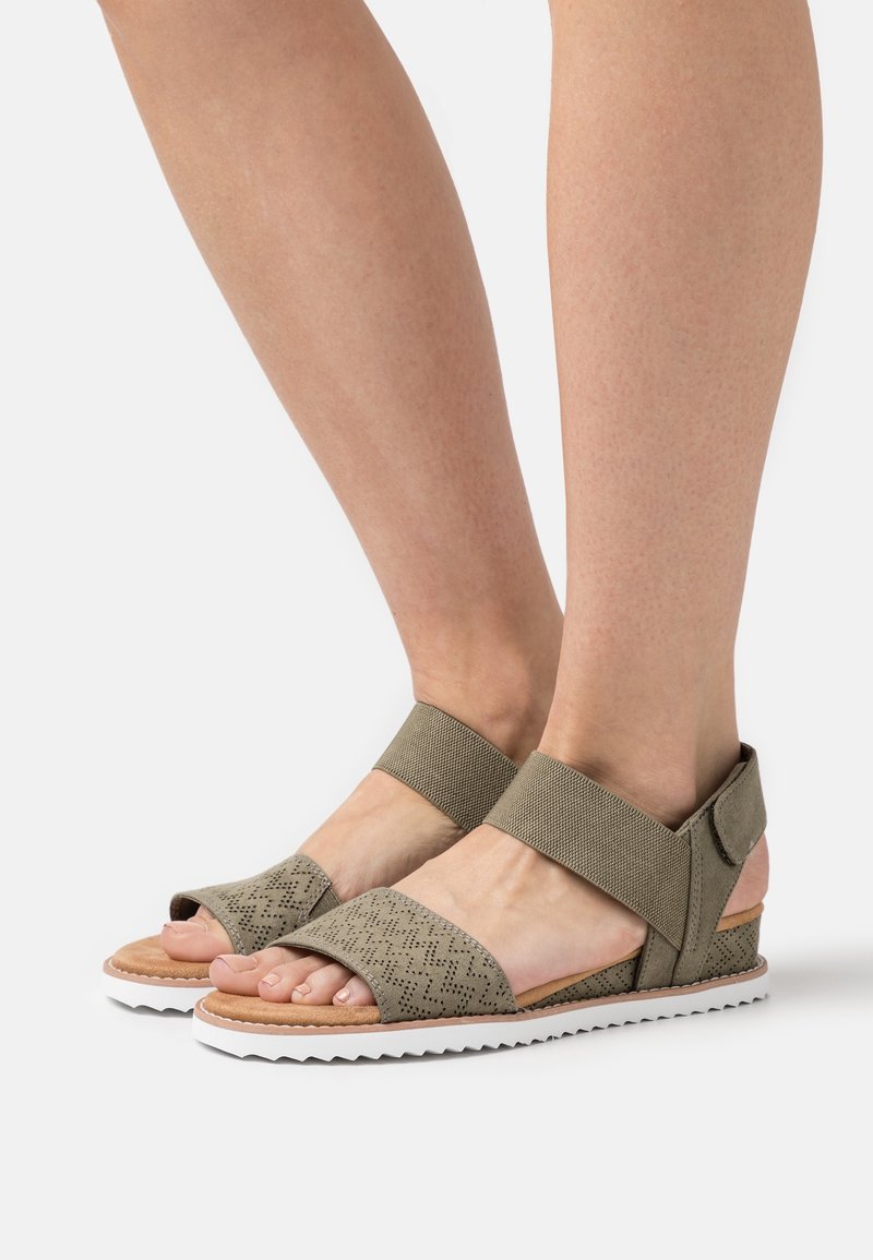 Pieds portant des sandales ouvertes beige avec des brides perforées en zigzag, des bandes élastiques à la cheville et des semelles blanches à rainures sur un fond blanc.