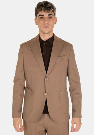Giovane uomo con capelli ricci corti che indossa un giubbotto di un completo beige e pantaloni abbinati sopra una camicia button-up marrone scuro, in piedi su uno sfondo bianco.