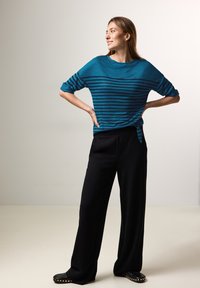 Turkoosisauva raitapaita käärittyillä hihoilla, yhdistettynä leveisiin mustiin housuihin ja mustiin kengissä, joissa on kultaiset niitit. Saumatonta kangasrakennetta.