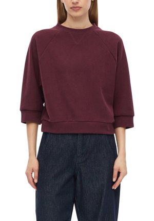 Frau trägt ein kastanienbraunes Sweatshirt mit Dreiviertelärmeln und dunkelblaue hochtaillierte Jeans vor weißem Hintergrund.