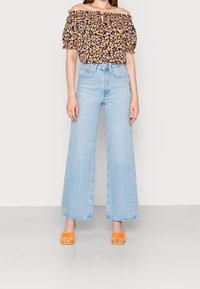 Haut noir à fleurs, épaules dénudées avec manches bouffantes, associé à un jean large bleu clair et des sandales ouvertes orange.