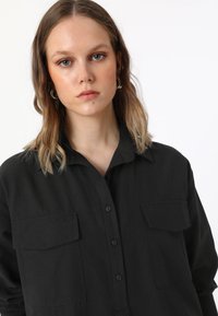 Chemise noire à boutons avec deux poches poitrine, manches longues et col. Le tissu semble lisse et léger. Design simple et fonctionnel.