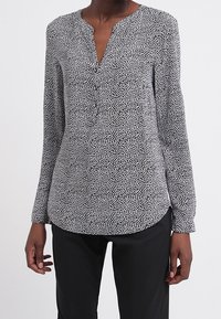 Vrouw in een zwart-witte blouse met lange mouwen en kleine abstracte vlekken, met zwarte broek, staand tegen een effen achtergrond.