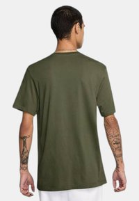 T-shirt di cotone verde oliva con vestibilità rilassata, maniche corte e collo girocollo. Presenta tatuaggi visibili sulle braccia.