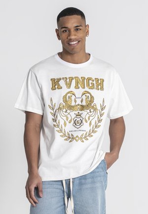 Jeune homme souriant, portant un t-shirt blanc avec un dessin de phénix doré et l'inscription "KVNGH", associé à un jean bleu clair, debout devant un fond gris.