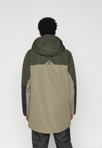 Kapuce turintis vandeniui atsparus džemperis, pasižymintis dviejų spalvų dizainu olivų žalia ir smėlio spalvos su tamsiai mėlynais akcentais. Pridedamas logotipo detalė viršutiniame nugaros regione.