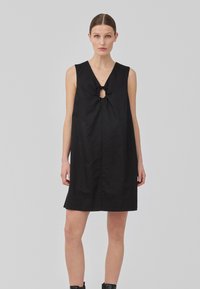 Robe sans manches noire avec un décolleté en V et une ouverture en forme de trou de serrure agrémentée d'un nœud. Le tissu semble lisse avec une silhouette droite et décontractée.